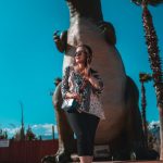 photo of a woman wearing a printed top and sunglasses standing near dinosaur figurine
